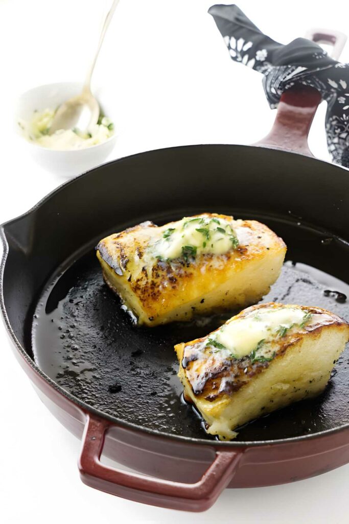 Two sea bass fillets with herb butter in a black skillet.