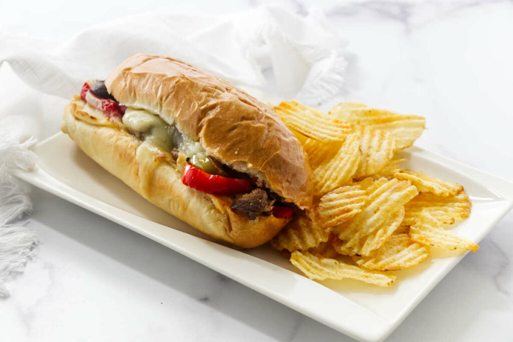 A philly cheesesteak on a plate with potato chips.