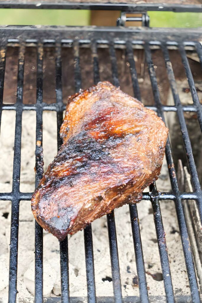 Seasoned tri tip roast grilling over charcoal with grill marks and ashy coals.