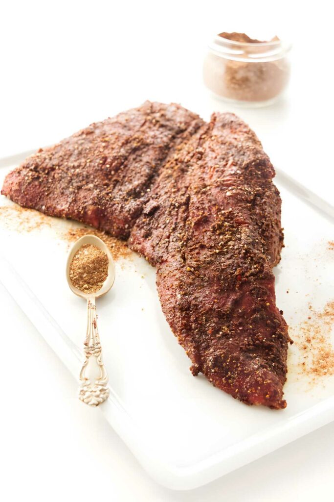Seasoned tri-tip steak on cutting board with dry rub and spice jar.