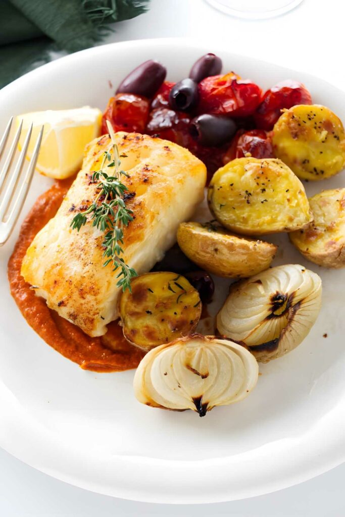 Baked white fish with herbs, roasted vegetables, red sauce, and lemon wedge.