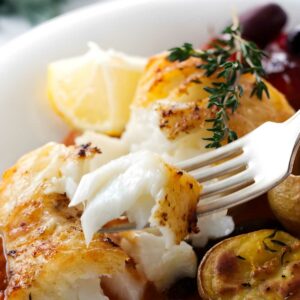 Fork cutting flaky baked fish fillet with thyme, potatoes, and lemon wedge.