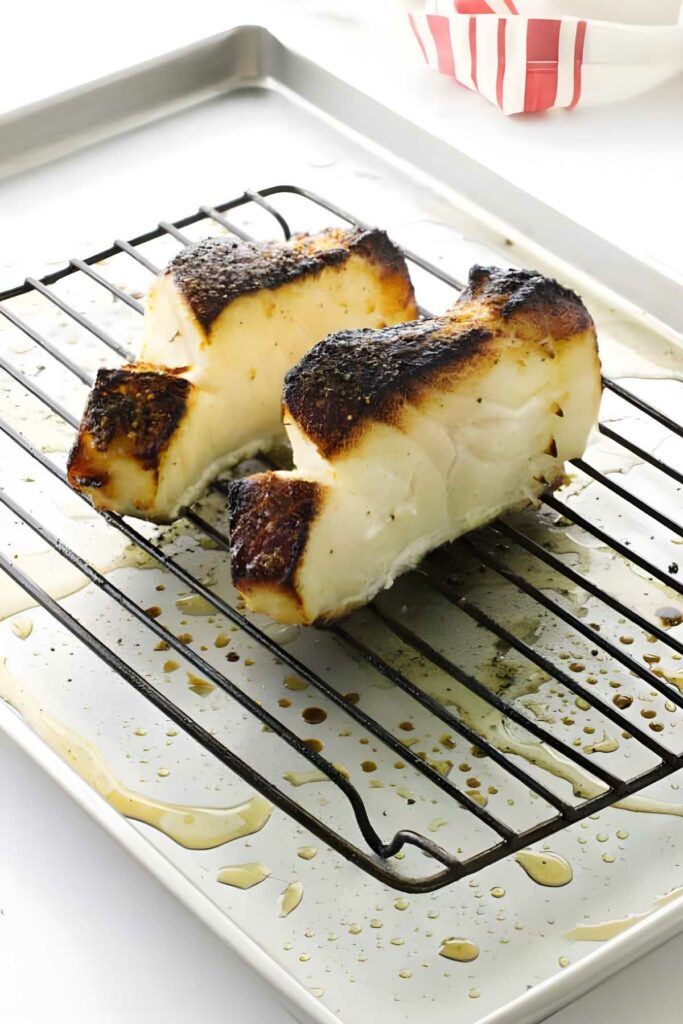 Two caramelized broiled sea bass fillets on rack over oily baking sheet.