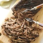 Cooked beef roast being shredded with forks on a cutting board.