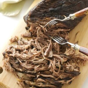 Cooked beef roast being shredded with forks on a cutting board.