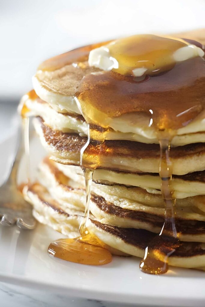Stack of ricotta sourdough pancakes with butter, syrup drizzling, and a fork on plate.