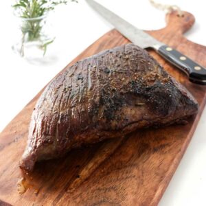Cooked beef on cutting board with knife; herbs in glass behind.