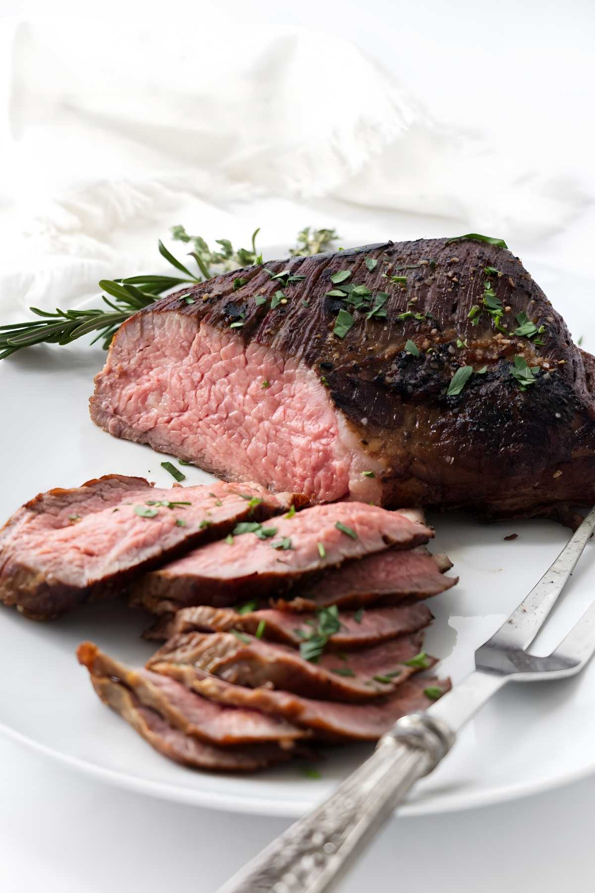 Sliced medium-rare tri tip with herbs and fork on white background.