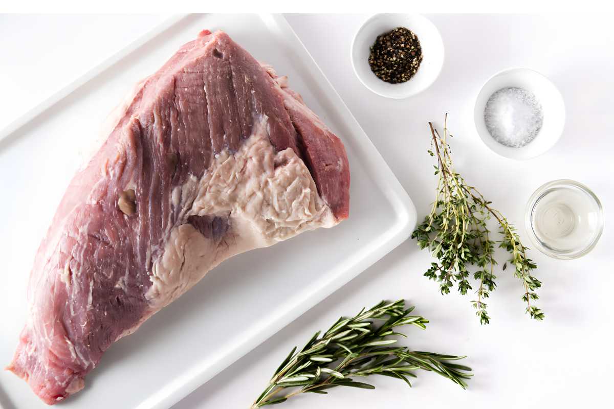 Raw tri tip roast with herbs, salt, pepper, and oil on white tray.