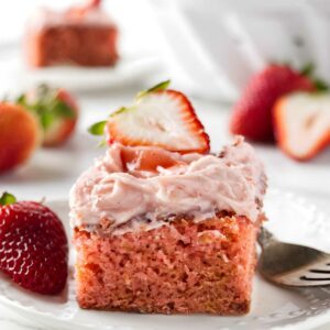 Strawberry cake slice with pink frosting and fresh strawberry on white plate.