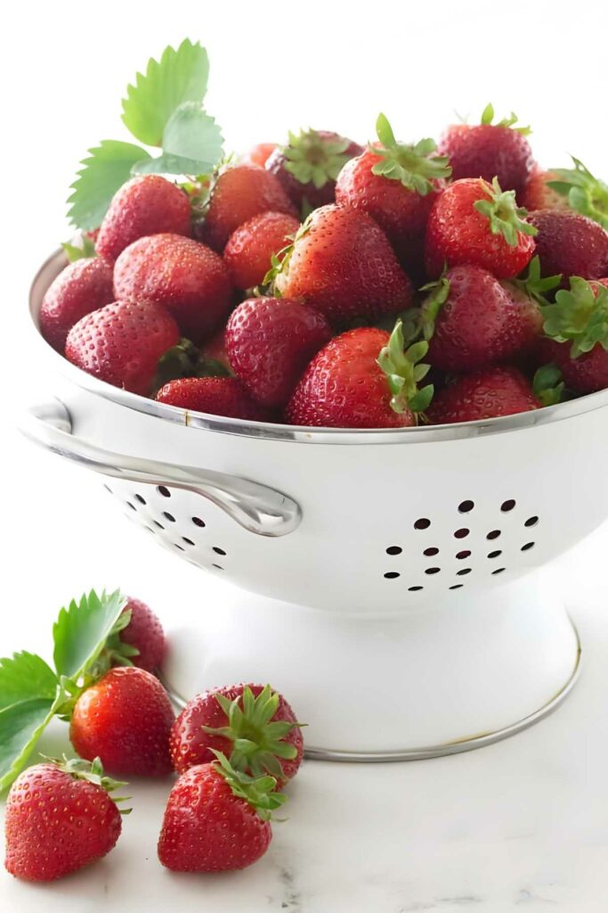 White colander of ripe strawberries with a few scattered on white surface.