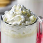Glass mug of white hot chocolate with whipped cream; cookies in background.