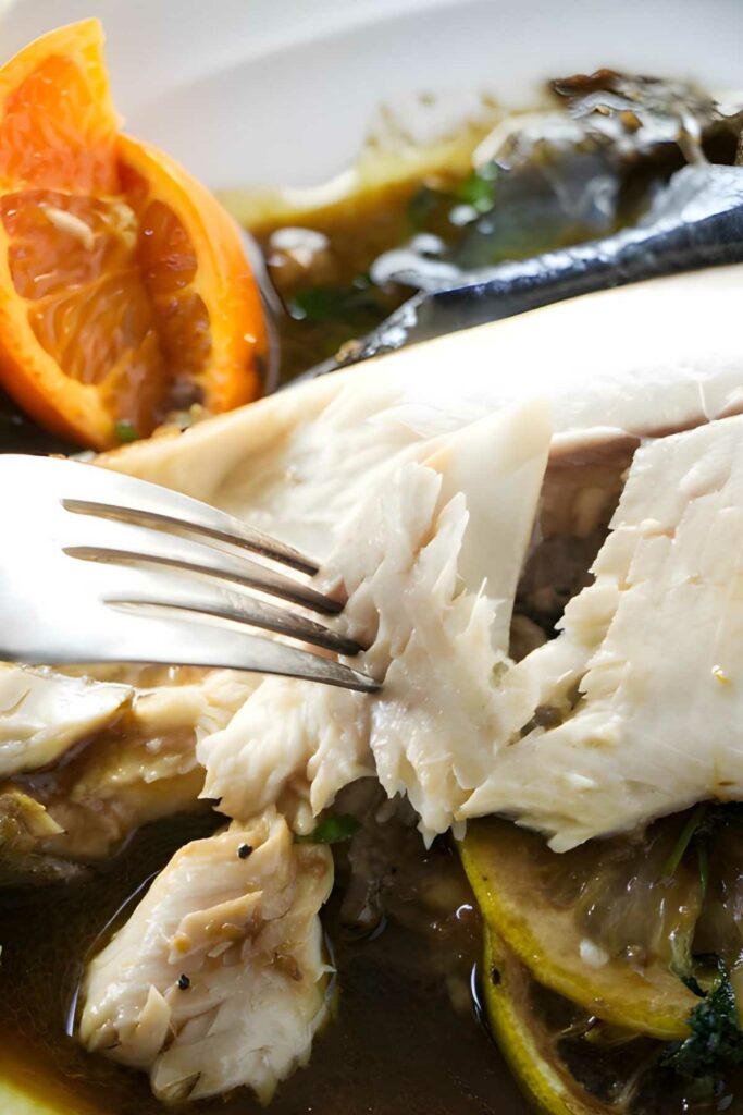 Fork flaking cooked white fish with lemon, orange, herbs, and broth.