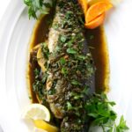 Whole cooked fish with herbs, citrus wedges, brown sauce, and parsley.