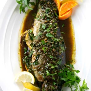 Whole cooked fish with herbs, citrus wedges, brown sauce, and parsley.