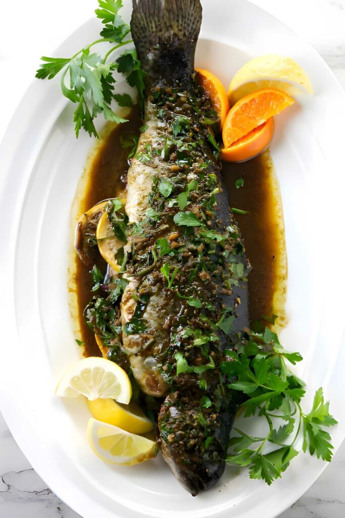 Whole cooked fish with herbs, citrus wedges, brown sauce, and parsley.