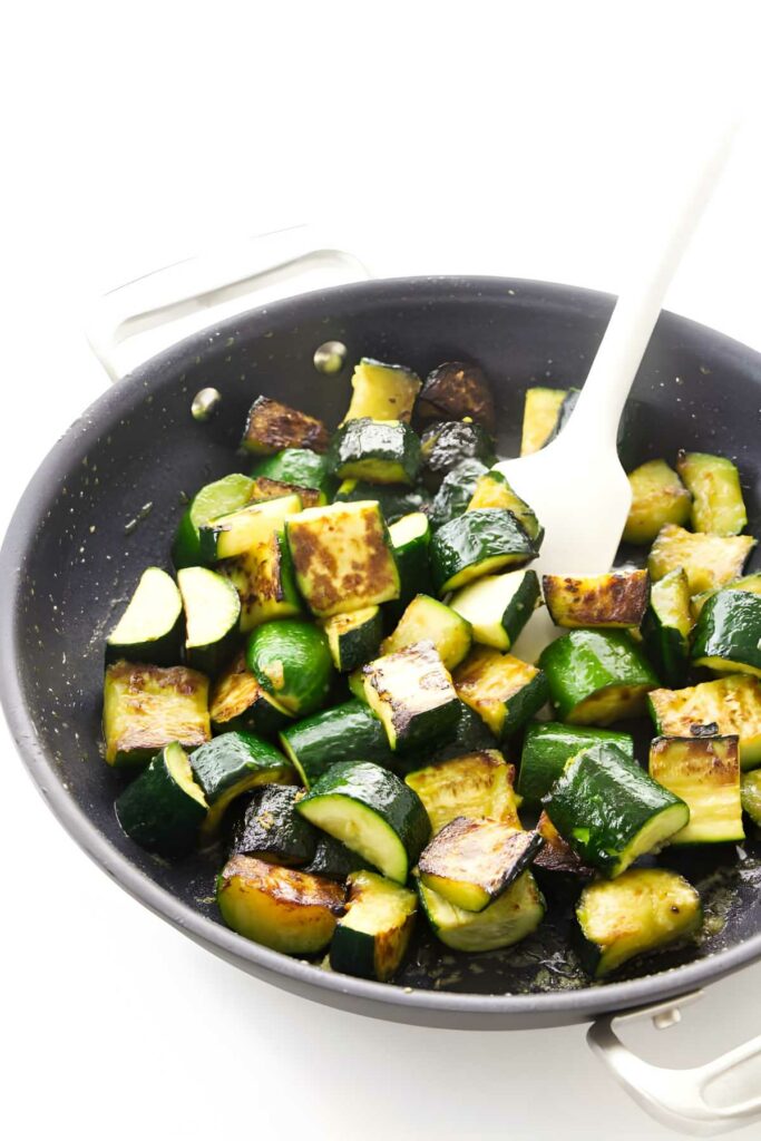 Chopped zucchini sautéing in black skillet, stirred with white spatula.