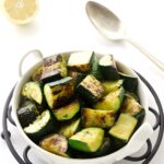 Bowl of grilled zucchini with spoon, lemon, and fresh herbs nearby.