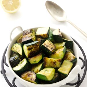 Bowl of grilled zucchini with spoon, lemon, and fresh herbs nearby.