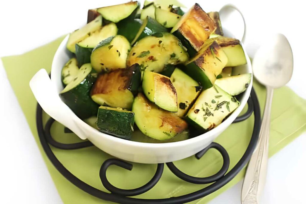 Bowl of grilled zucchini chunks with herbs on green napkin.