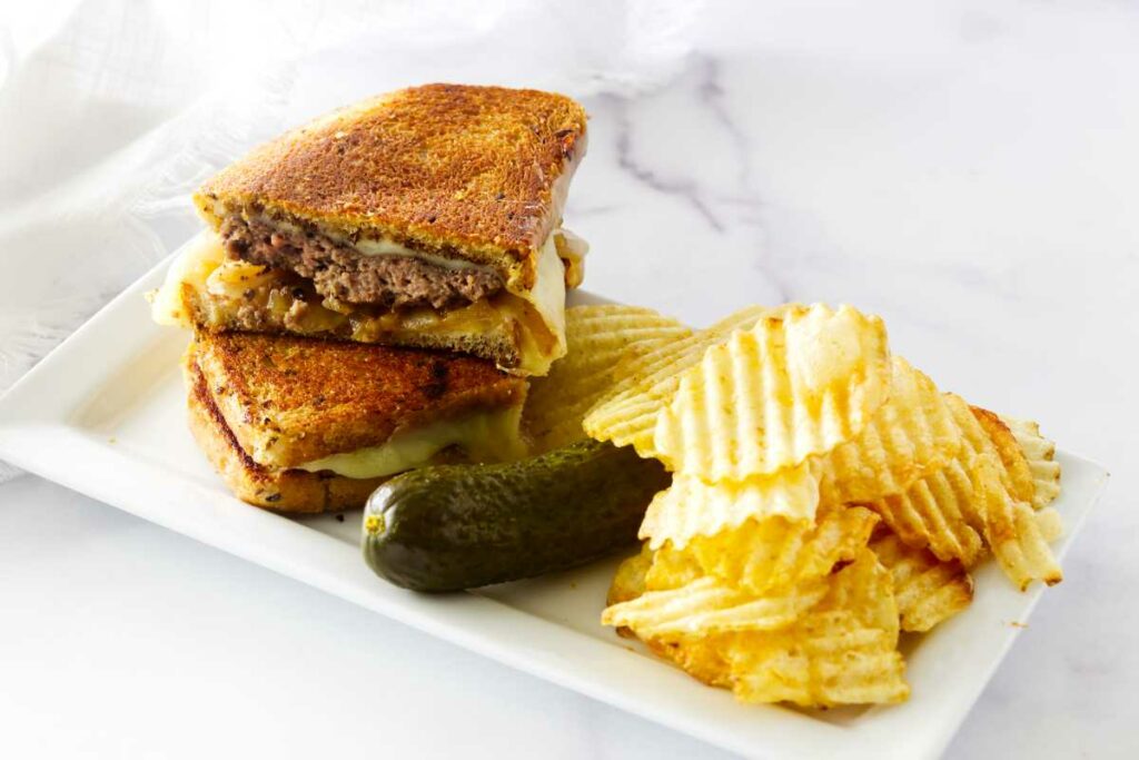 A skillet diner-style patty melt on a plate with potato chips and a pickle.