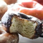 Close-up of bitten chocolate donut atop another, with blurred donuts behind.