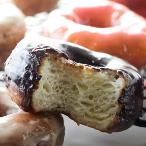 Close-up of bitten chocolate donut atop another, with blurred donuts behind.