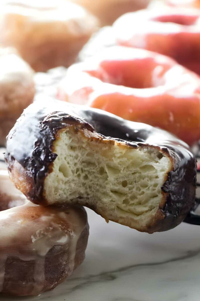 Close-up of bitten chocolate donut atop another, with blurred donuts behind.