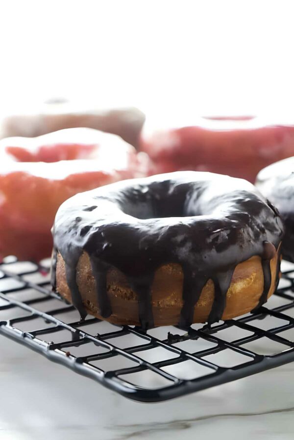 Chocolate-glazed donut on cooling rack, pink donuts blurred in background.