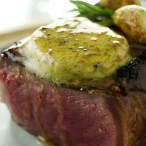 Medium-rare steak with melting herb butter, garnished with herbs and potatoes.