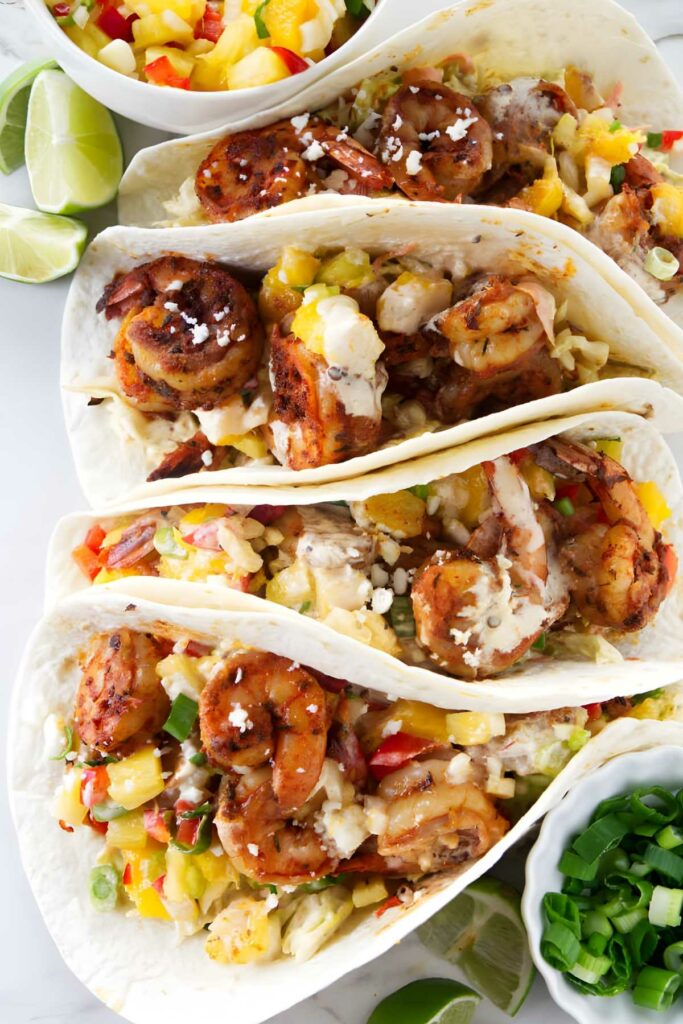 Three spicy shrimp tacos with mango salsa, cheese, limes, and green onions.