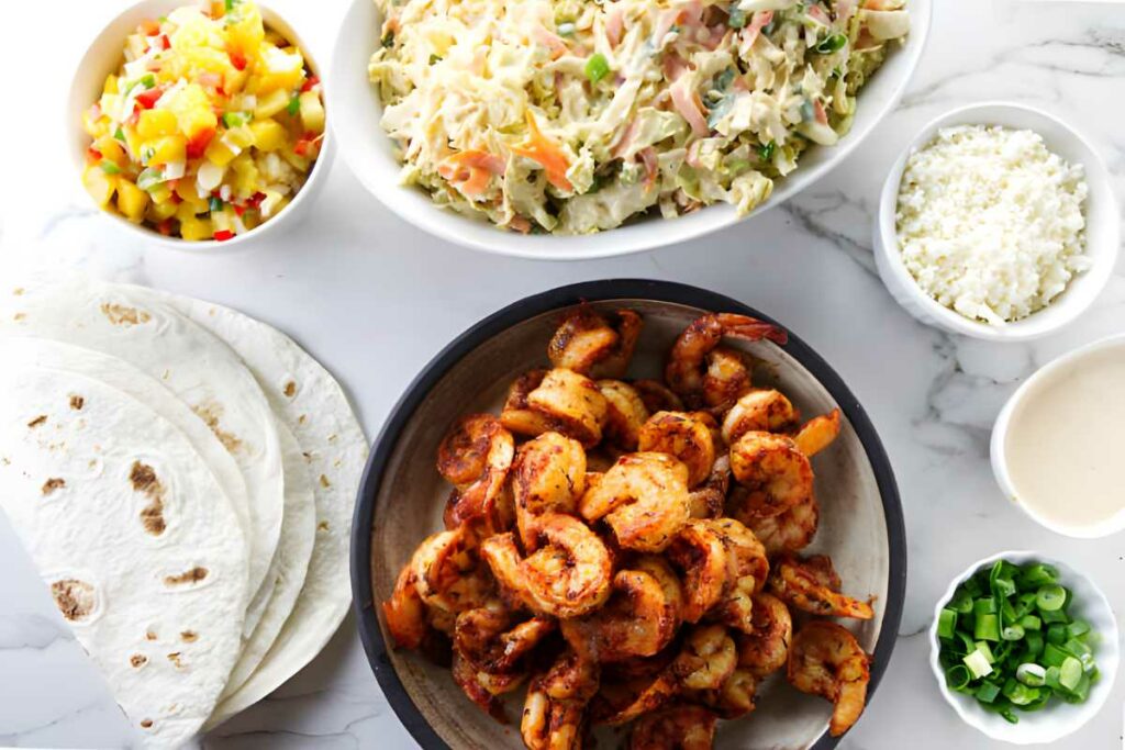 Shrimp taco ingredients spread on white surface, including shrimp, tortillas, and toppings.