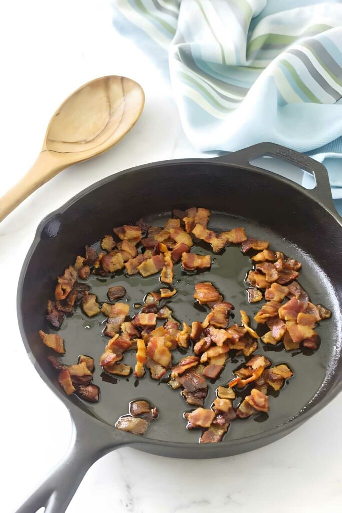 Black skillet with cooked bacon, wooden spoon, and blue-striped cloth nearby.