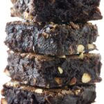 Stack of four fudgy brownies with nut chunks on white surface.