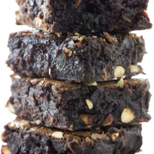 Stack of four fudgy brownies with nut chunks on white surface.