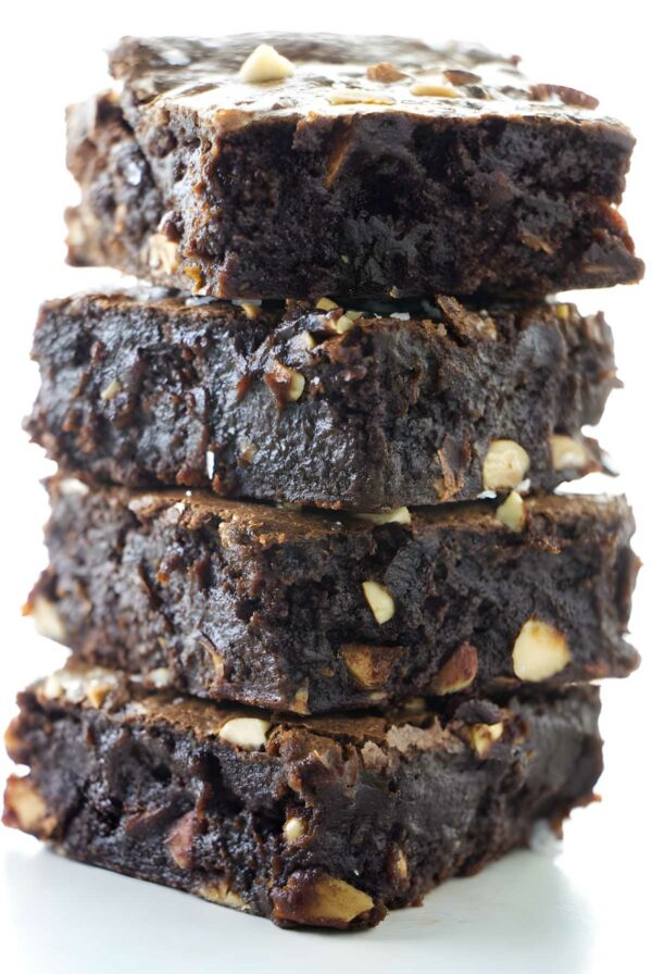 Stack of four fudgy brownies with nut chunks on white surface.