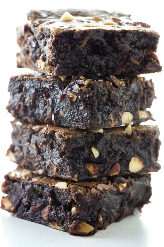 Stack of four fudgy brownies with nut chunks on white surface.
