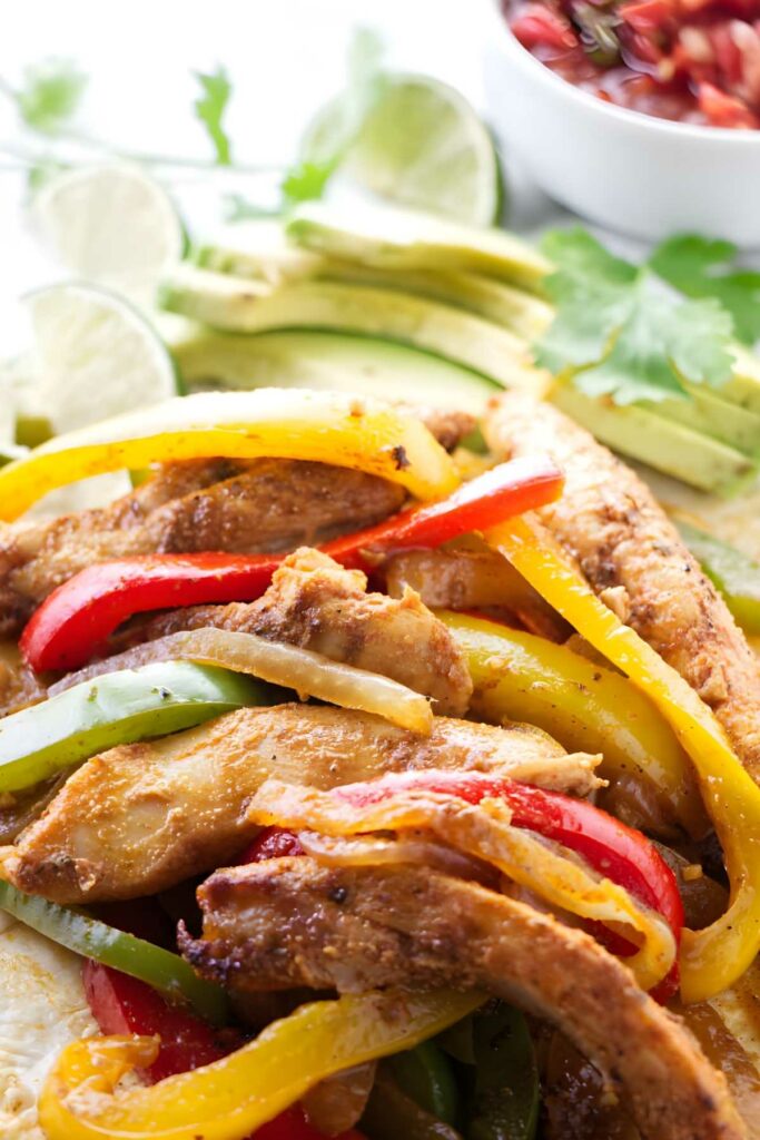 Chicken fajitas with grilled peppers, avocado slices, lime wedges, and cilantro.
