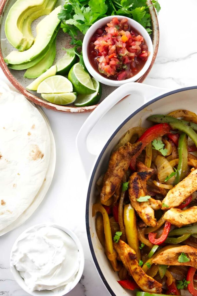 Bowl of chicken fajitas with peppers, tortillas, avocado, salsa, and sour cream.