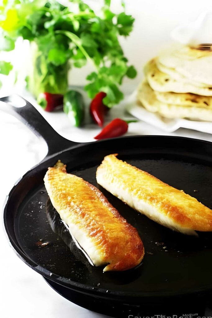 Two tilapia fish fillets cooking in skillet, fresh herbs and flatbreads behind.