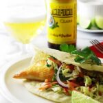 Fish taco with slaw and lime, beer and bottle in background.