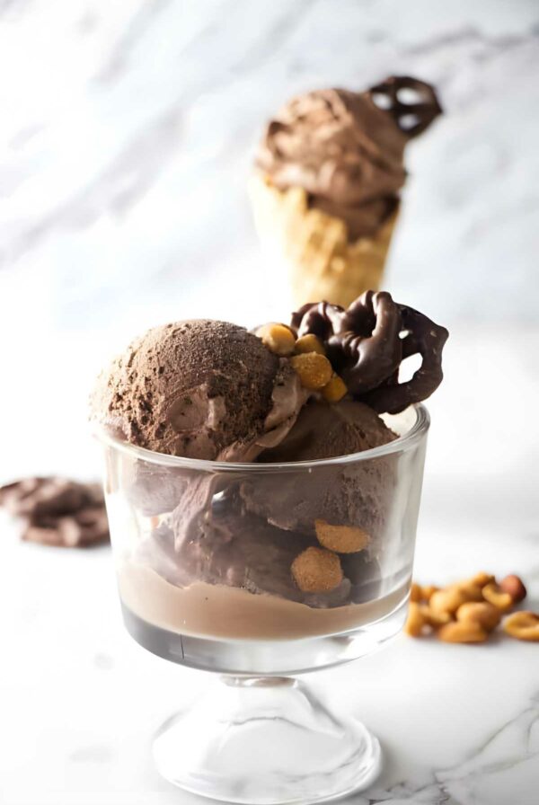 Glass dish of chocolate ice cream with pretzels, nuts; cone in background.