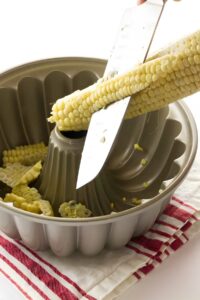 Person cutting corn off cob into Bundt pan on striped towel.