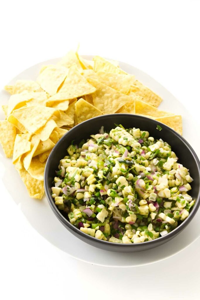 Black bowl of corn salsa with tortilla chips on a white plate.