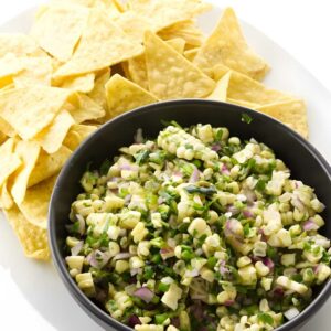 Black bowl of corn salsa with chips on a white plate.