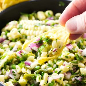 Hand holding tortilla chip with colorful corn salsa over bowl, chips behind.