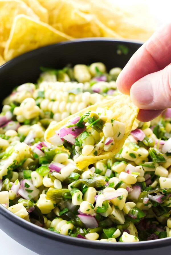 Hand holding tortilla chip with colorful corn salsa over bowl, chips behind.