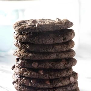 Stack of thin chocolate cookies on marble, blurred cookies behind.