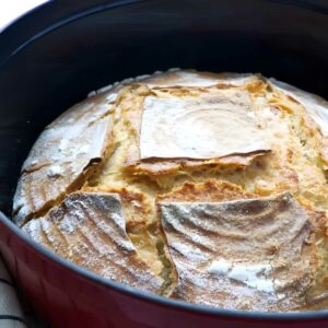 Golden-brown crusty loaf in red Dutch oven on white background.