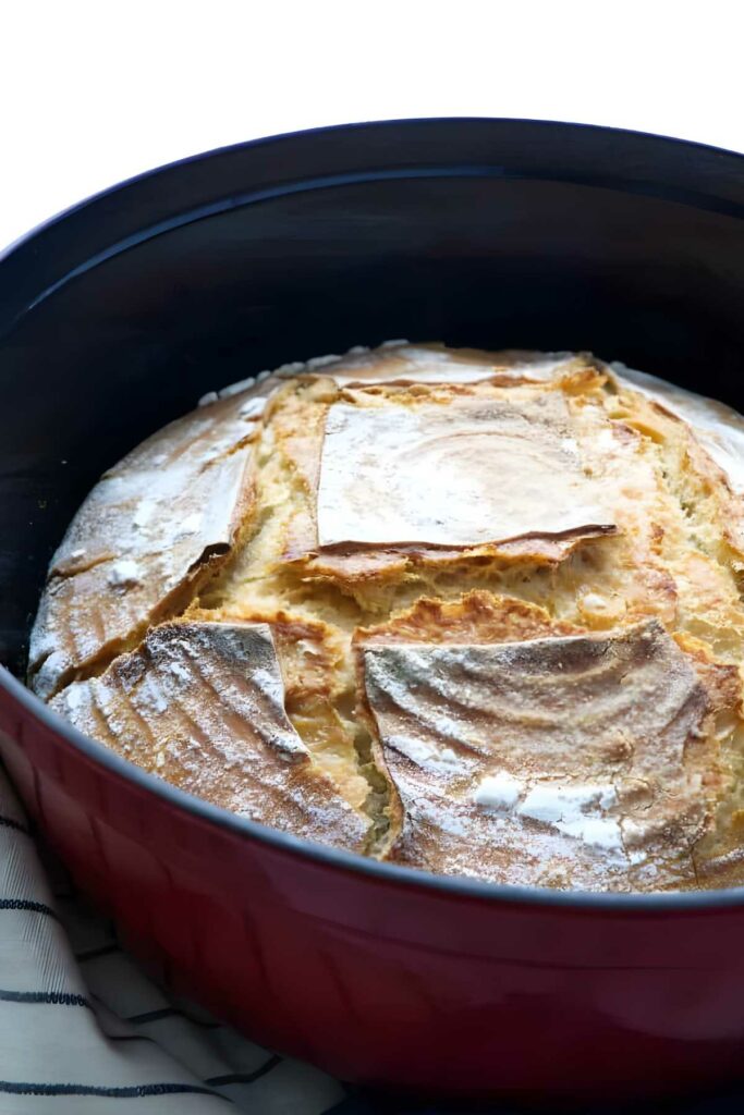 Golden-brown crusty loaf in red Dutch oven on white background.